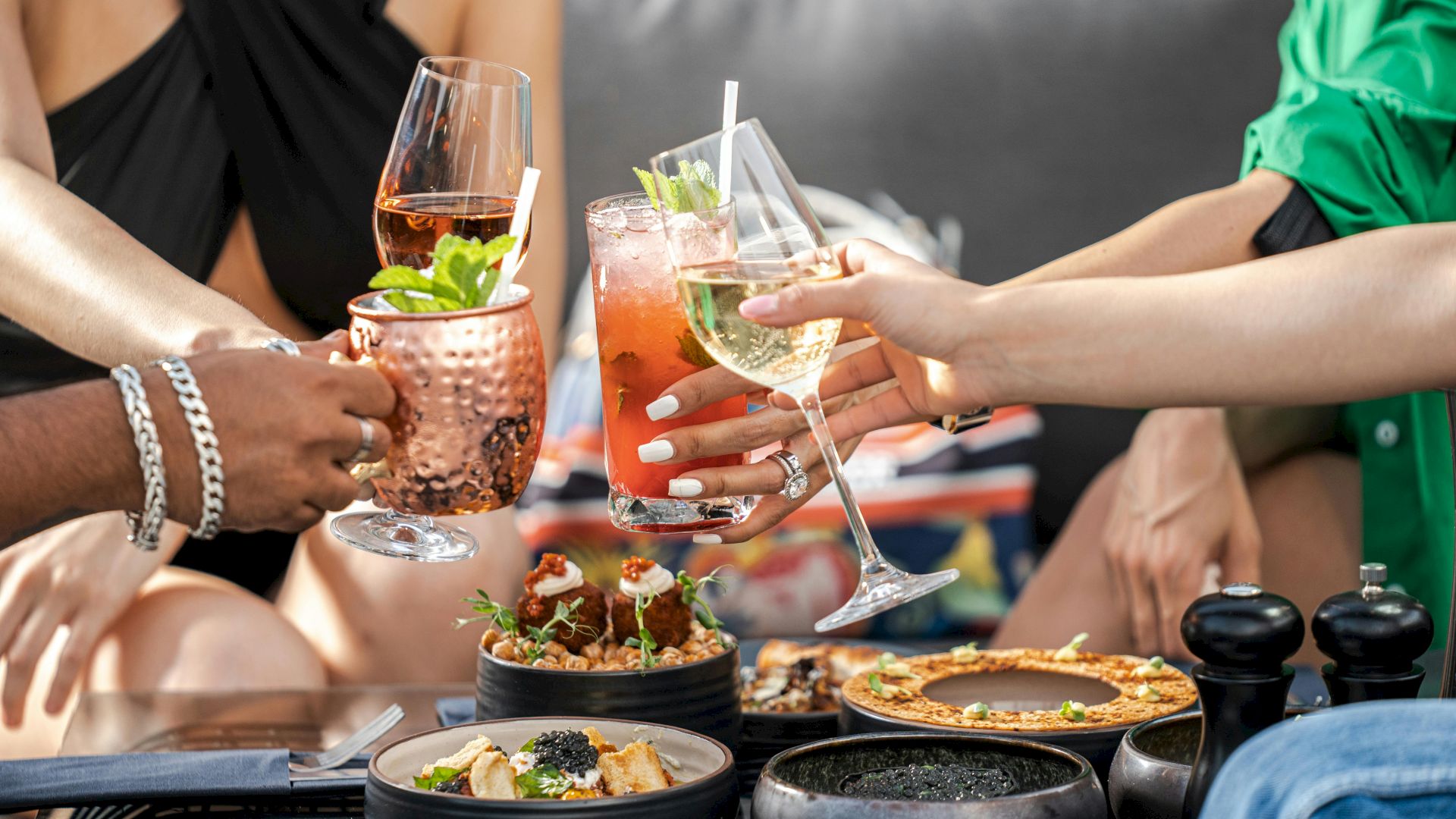 A group of friends cheers with assorted cocktails over a table of tapas and small plates at a lively outdoor get-together.