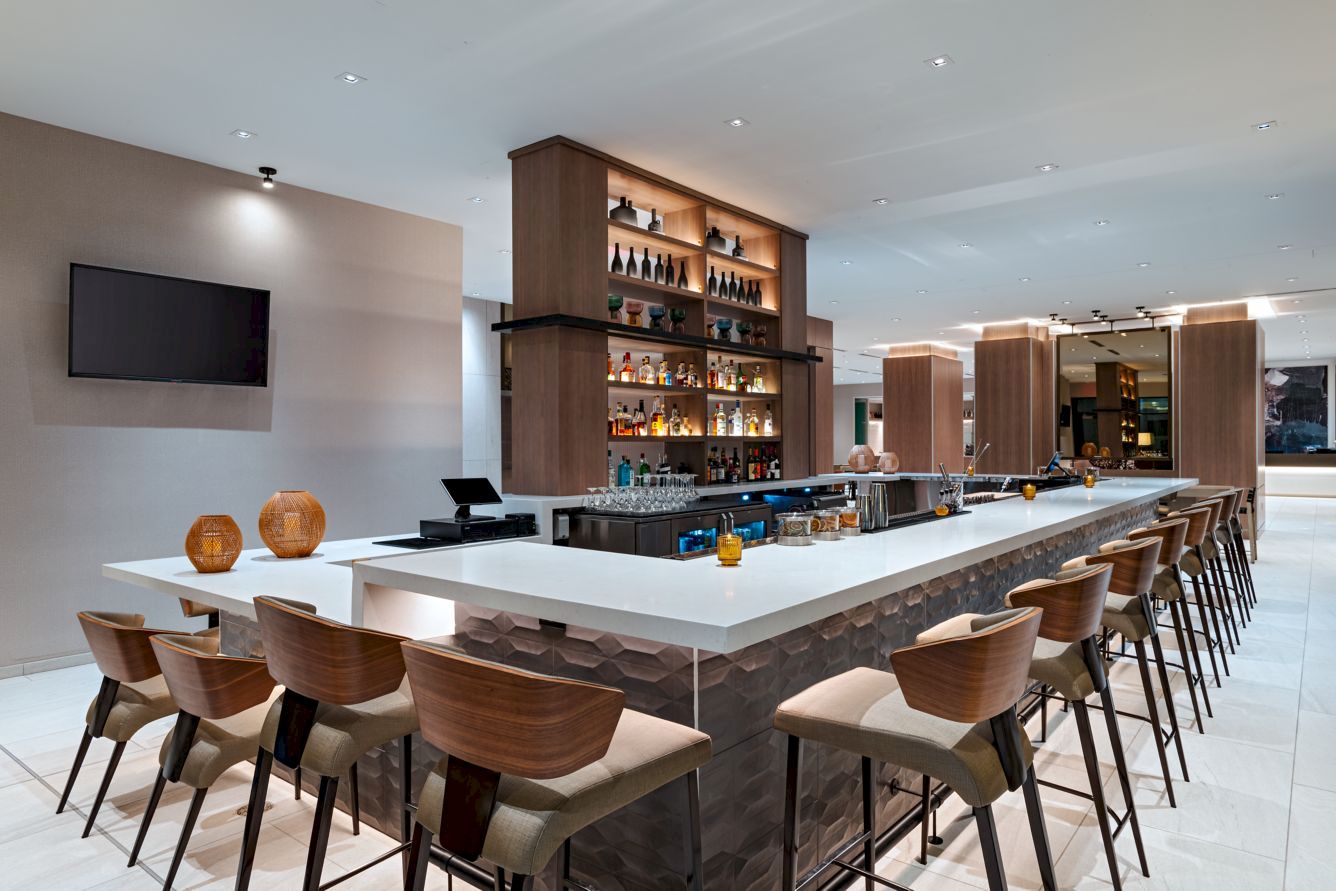 A modern hotel bar with a long white-topped island, wooden stools, shelving stocked with bottles, and a wall-mounted TV in a bright, open space. The bar area is lit warmly.