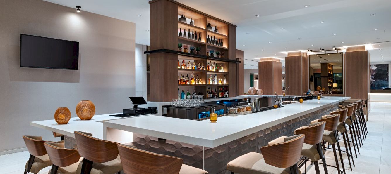 A modern hotel bar with a long white counter, wooden stools, a backlit shelf of liquor, and a sleek, well-lit lounge area.