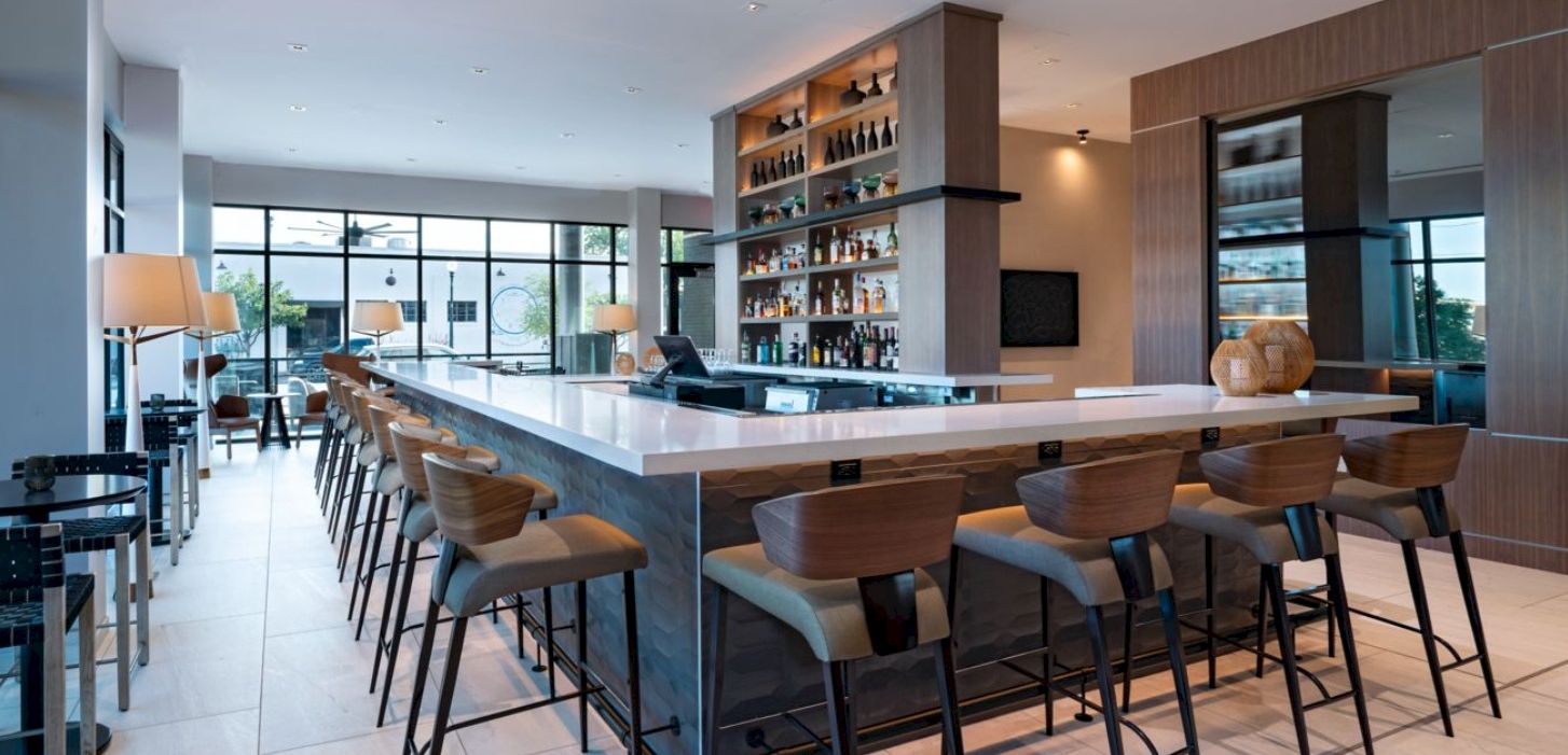 A modern, airy hotel bar with a long white counter, wooden stools, and a well-stocked backlit shelf, overlooking a sunny lounge area.