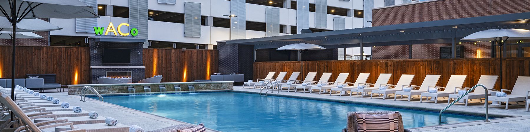 A modern hotel pool area with lounge chairs, a fire pit, and a bright building backdrop at dusk, inviting and stylish.