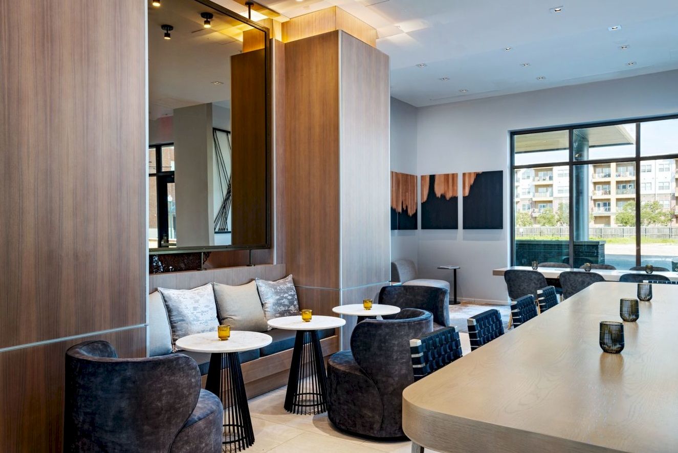 A modern lounge with a curved bar, dark chairs, small round white tables, and large windows offering a sunny city view.