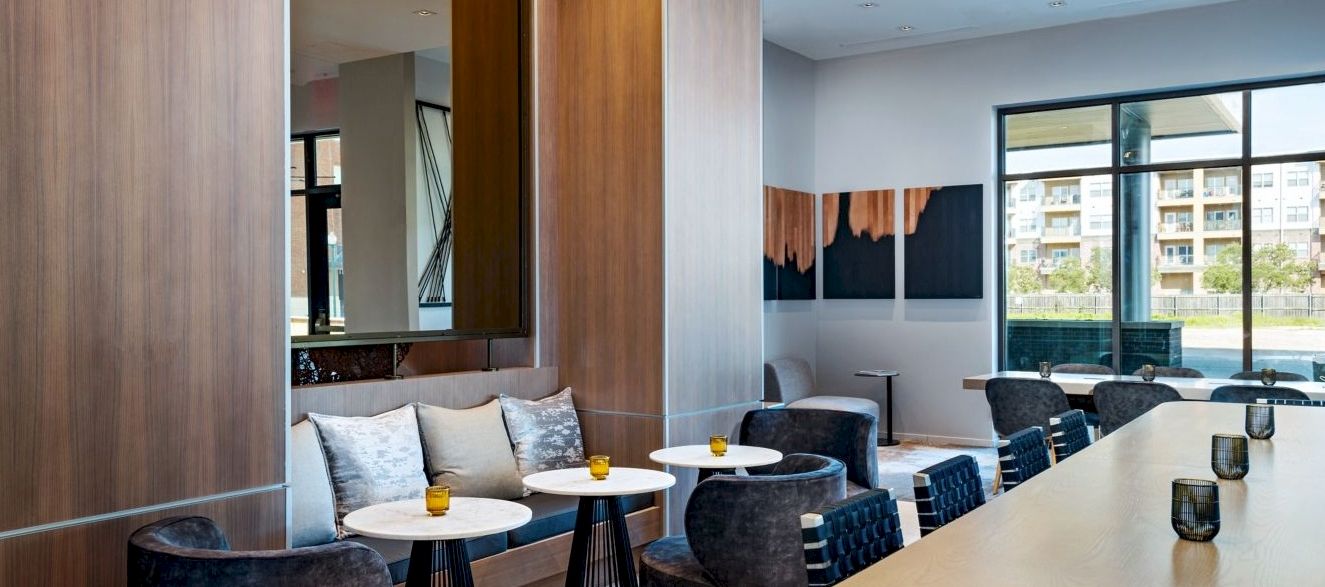 A modern lounge with a curved bar, dark chairs, small round white tables, and large windows offering a sunny city view.