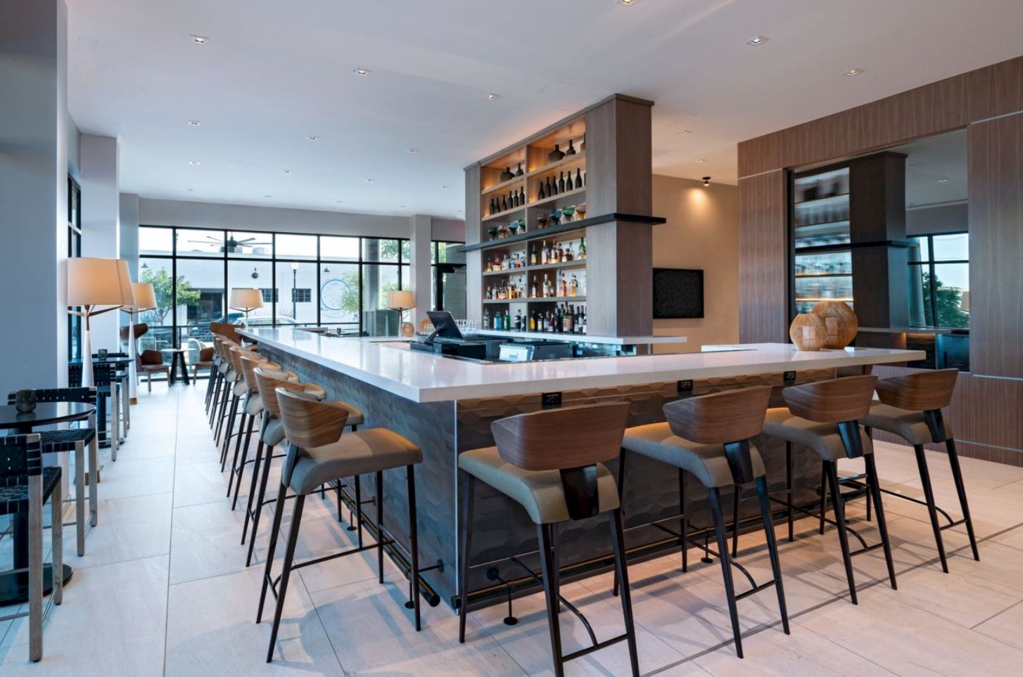 A modern hotel bar with a large white island, wooden stools, tall shelves of bottles, ambient lighting, and a bright lounge area in the background.
