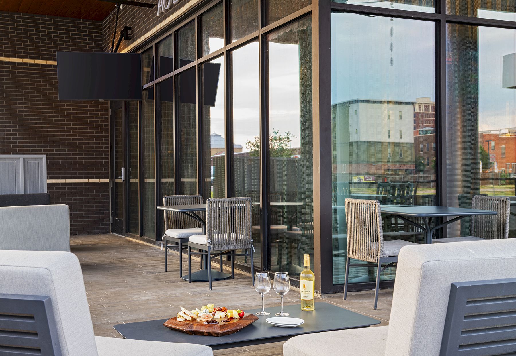A stylish outdoor dining area with glass walls, modern chairs, a small table set with wine, two glasses, and a plate of appetizers, ready for a sunset meal.