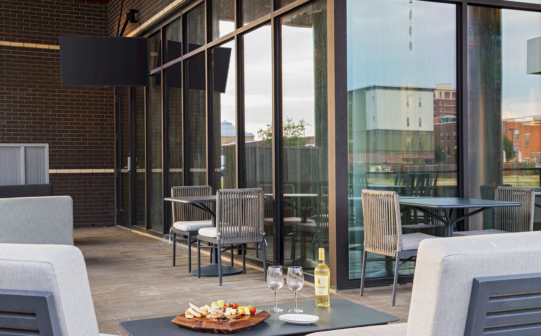 A stylish outdoor dining area with glass walls, modern chairs, a small table set with wine, two glasses, and a plate of appetizers, ready for a sunset meal.