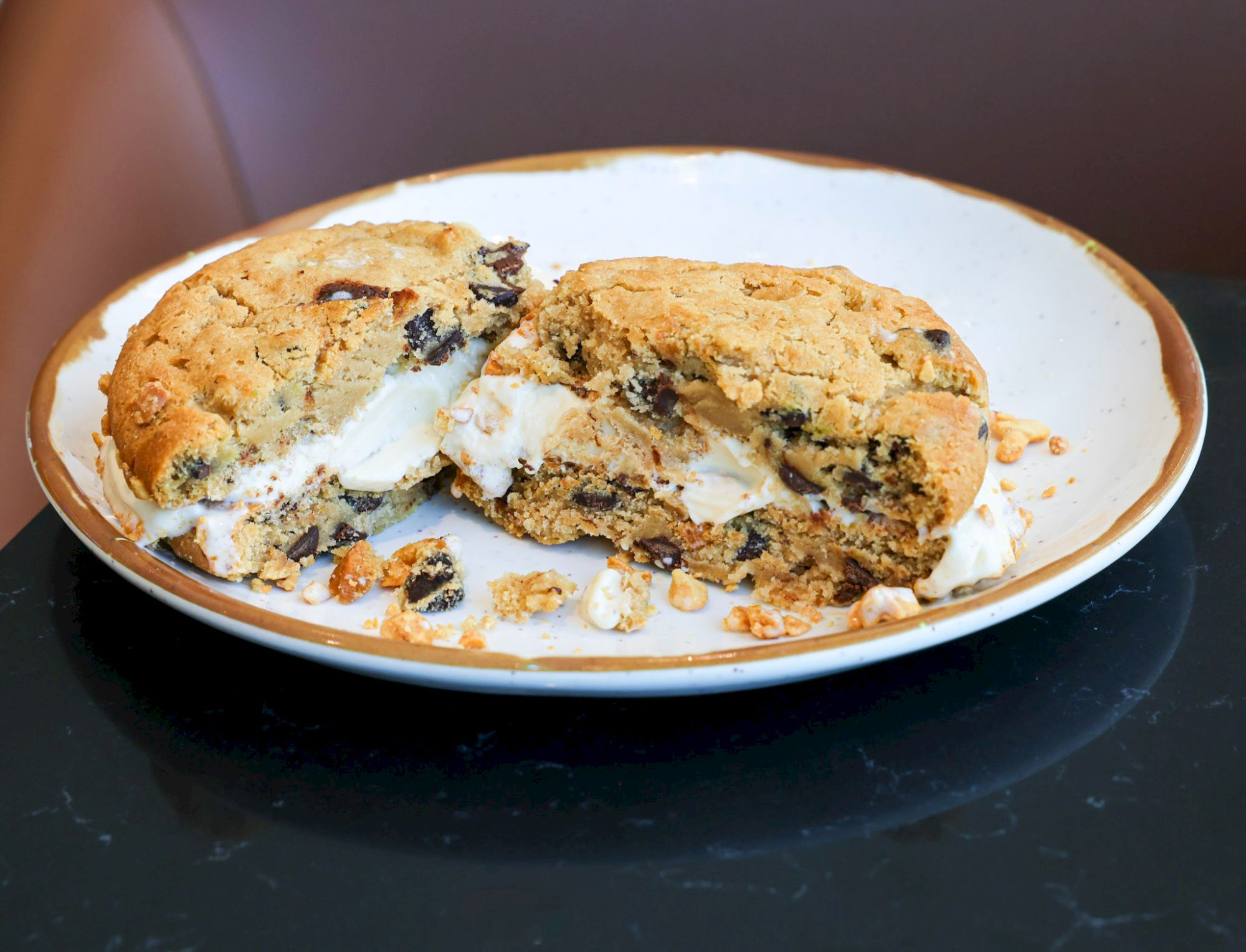 Two broken cookies with vanilla ice cream melting inside on a white plate with a brown rim.