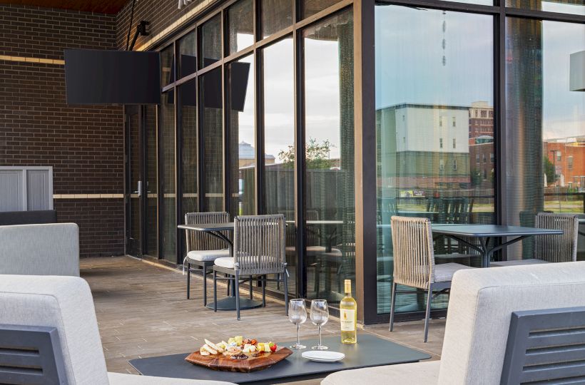 A stylish outdoor dining setup with two gray couches, a small table, wine bottle, and glasses on a plate of snacks, under a glass-walled patio.