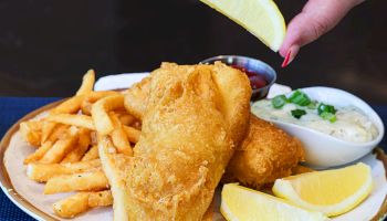 A plate with fried fish, french fries, lemon wedges, and a side of tartar sauce; a hand squeezes a lemon over the fish.