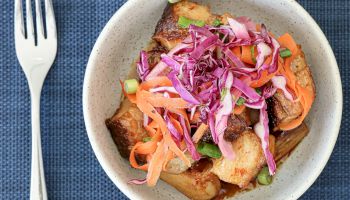 A bowl of roasted potatoes topped with colorful pickled cabbage and carrot slaw, served with a fork on a blue placemat.