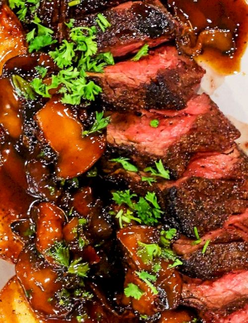 A sliced steak with rich brown gravy, saut&eacute;ed mushrooms/onions, and chopped parsley on a white plate.