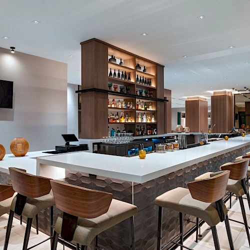 A modern hotel bar with a long white-topped island, wooden chairs, backlit shelves of liquor, a wall-mounted TV, and a bright, airy lounge area.