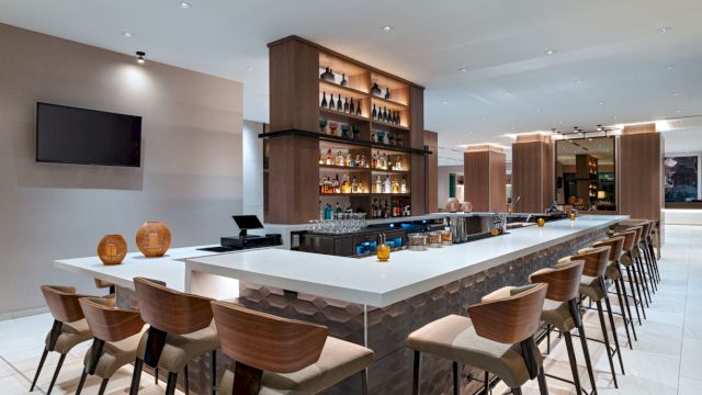 A modern hotel bar with a long white-topped island, wooden chairs, backlit shelves of liquor, a wall-mounted TV, and a bright, airy lounge area.