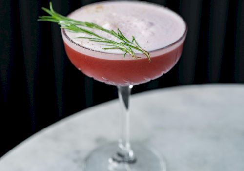 A pink cocktail in a coupe glass with a rosemary sprig garnish on top, set against a dark background.