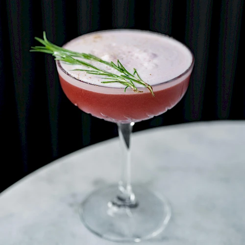A pink cocktail in a coupe glass with a rosemary sprig garnish on top, set against a dark background.