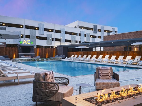 A modern hotel pool area with lounging chairs around a rectangular pool, fire pit seating in front, and a multi-story building backdrop at dusk.