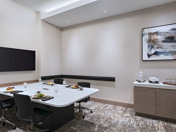 A modern conference room with a white table, six black chairs, a wall-mounted TV, a sideboard with snacks, and abstract wall art, all in neutral tones.