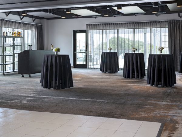 A modern event lounge with long cocktail tables draped in black, floral centerpieces, a bar cart, and large floor-to-ceiling windows.