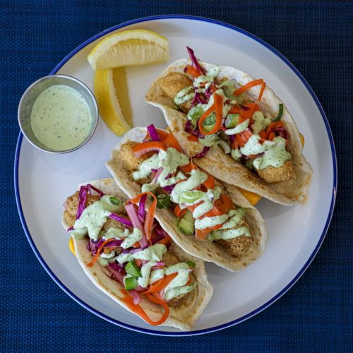 A plate of two small tacos or quesadillas topped with bacon bits and sauce, served with lime wedges and a dipping sauce on the side.