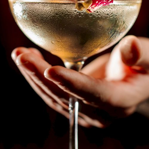 A close-up of a hand holding a cocktail glass, with a pale drink and a red garnish.