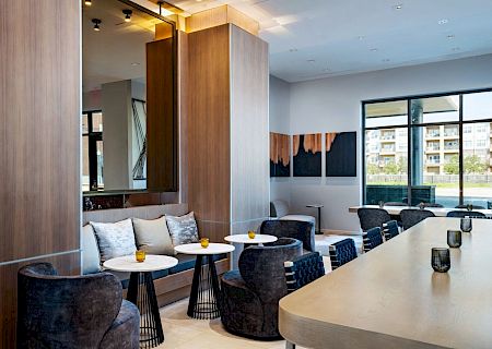 A modern lounge area with wood-paneled walls, a long light counter, and small round tables with dark chairs; large windows let in natural light.