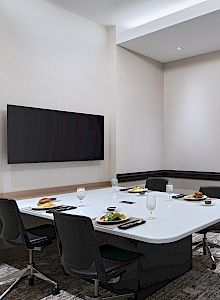 A modern conference room with a white table, black chairs, a wall-mounted TV, a sideboard with snacks, and abstract wall art, well lit.