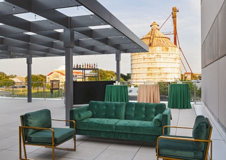An outdoor patio lounge with green sofas and tables beside a towering orange industrial furnace stack.