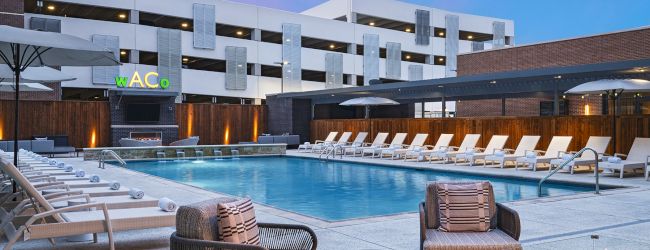 A luxury outdoor hotel pool area with lounge chairs, wicker seating, a fire pit, and modern white buildings in the background, under a blue sky.