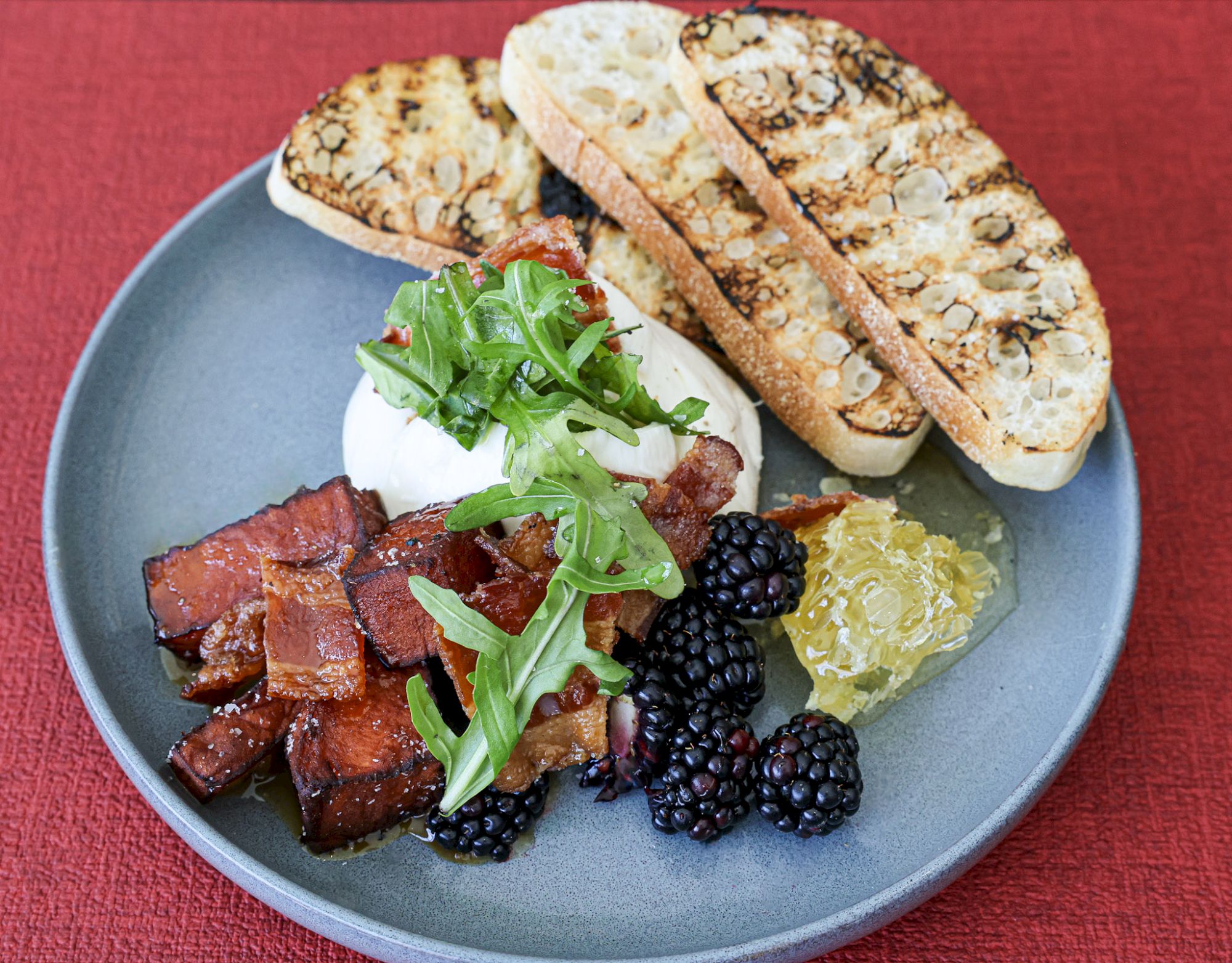 Cheesy bruschetta with grilled bread, crisp bacon-style meat, arugula, blackberries, and a dollop of relish on a blue plate.