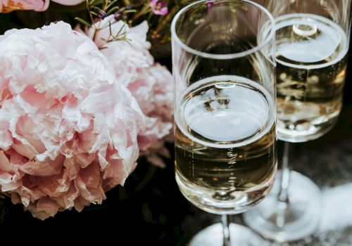 Two glasses of white wine beside soft pink peonies and delicate flowers arranged on a reflective table.