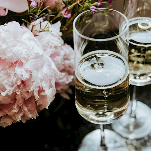Two glasses of white wine beside soft pink peonies and delicate flowers arranged on a reflective table.