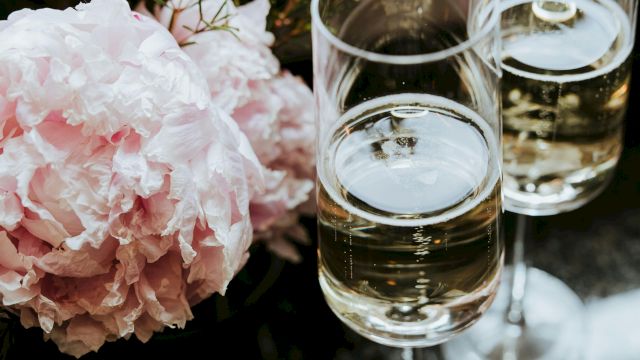 Two glasses of white wine beside soft pink peonies and delicate flowers arranged on a reflective table.