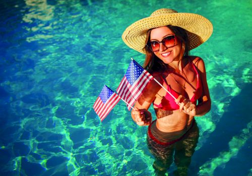 A woman in a straw hat and sunglasses stands in a clear blue pool, wearing a red bikini and holding small American flags.