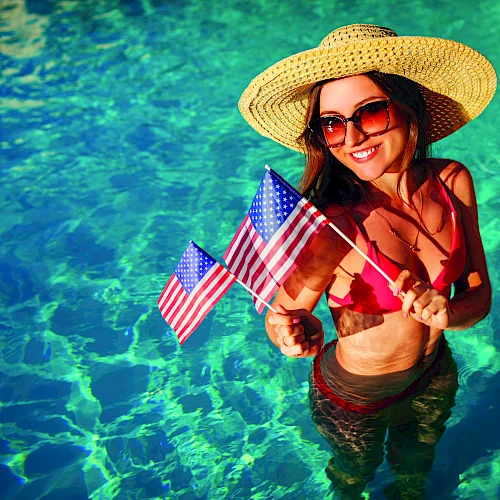 A woman in a straw hat and sunglasses stands in a clear blue pool, wearing a red bikini and holding small American flags.