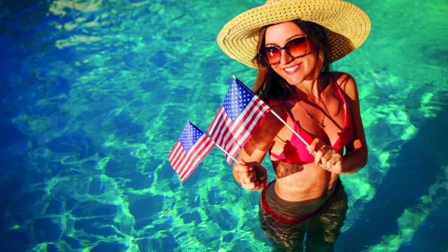 A woman in a straw hat and sunglasses stands in a clear blue pool, wearing a red bikini and holding small American flags.