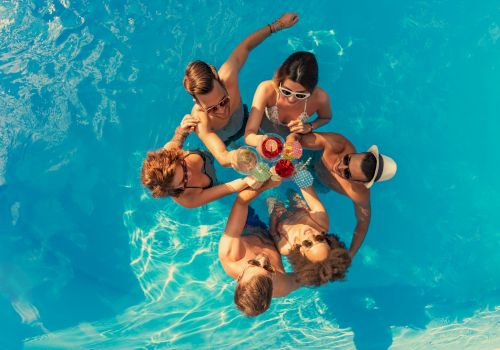 A group of friends are gathered in a bright blue pool, huddled for a toast with tropical drinks.