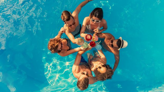 A group of friends are gathered in a bright blue pool, huddled for a toast with tropical drinks.