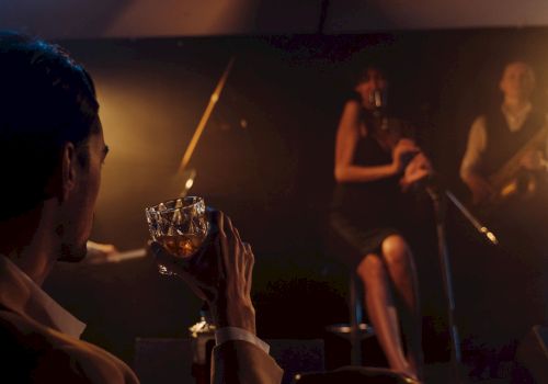 A live band performs in a dim, warm-lit venue while a person in the foreground holds a drink near the stage.