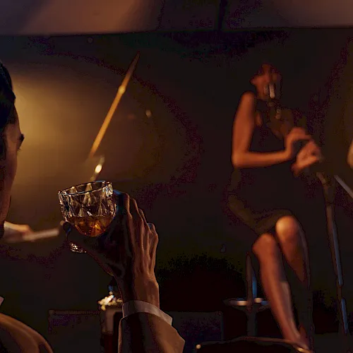 A live band performs in a dim, warm-lit venue while a person in the foreground holds a drink near the stage.