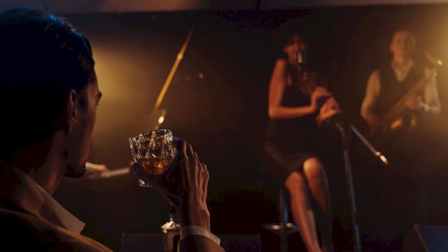 A live band performs in a dim, warm-lit venue while a person in the foreground holds a drink near the stage.