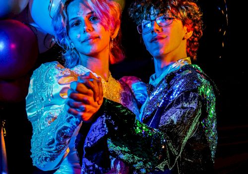 Two dancers in glittering show outfits pose under colorful stage lights, their faces highlighted in blue and orange glow.