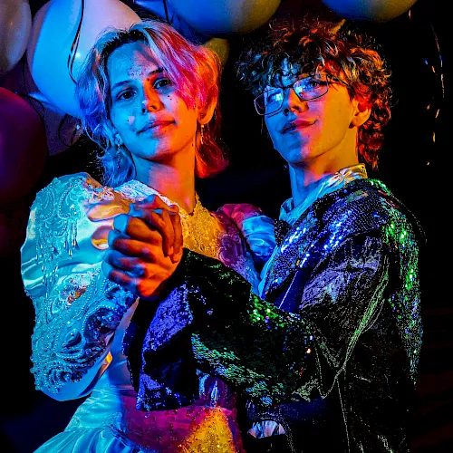 Two dancers in glittering show outfits pose under colorful stage lights, their faces highlighted in blue and orange glow.