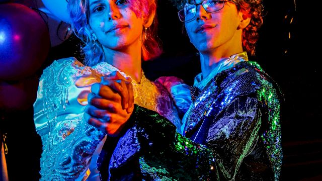 Two dancers in glittering show outfits pose under colorful stage lights, their faces highlighted in blue and orange glow.