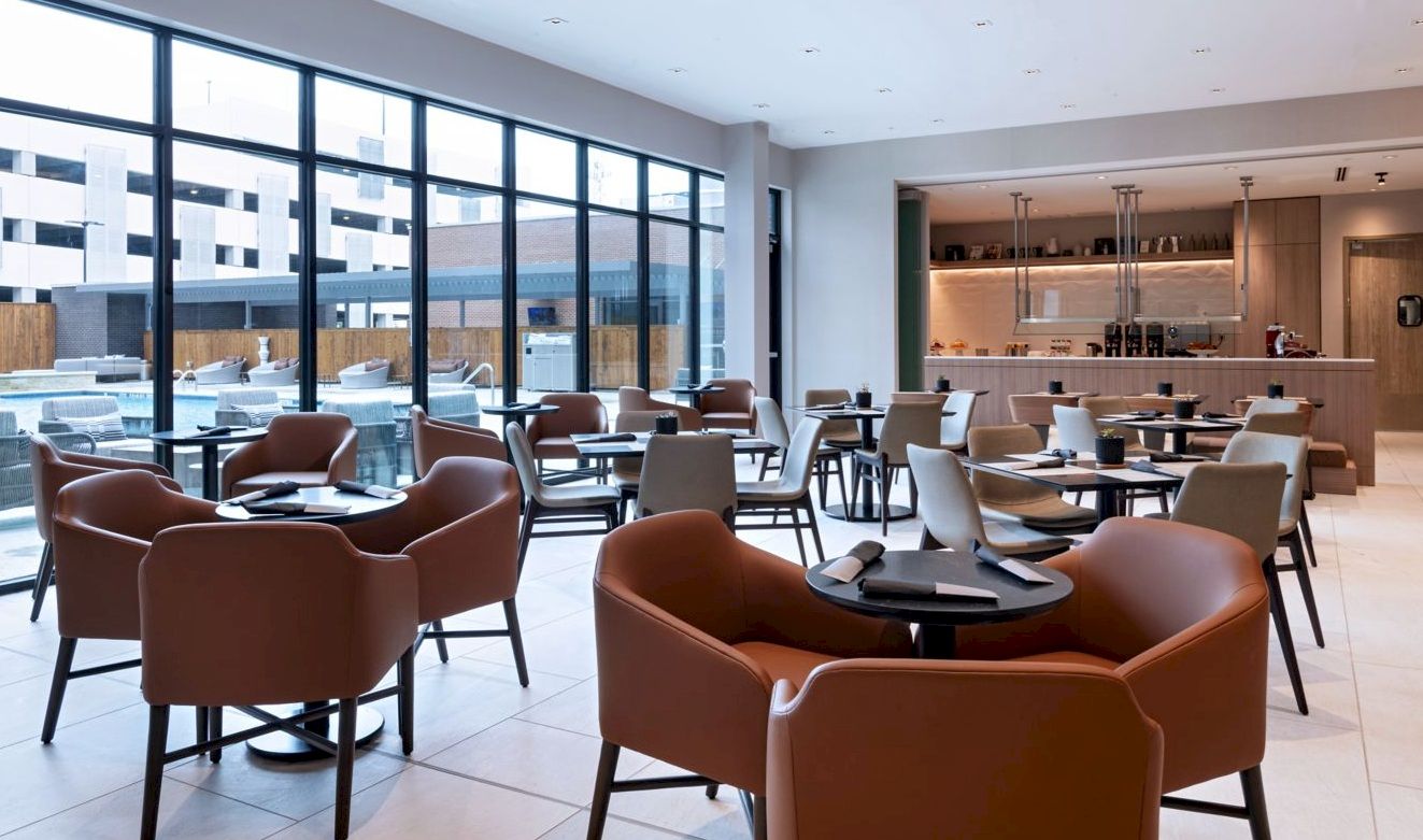 A modern office lounge/dining area with brown armchairs, round tables, large windows, and an open kitchenette in the back.
