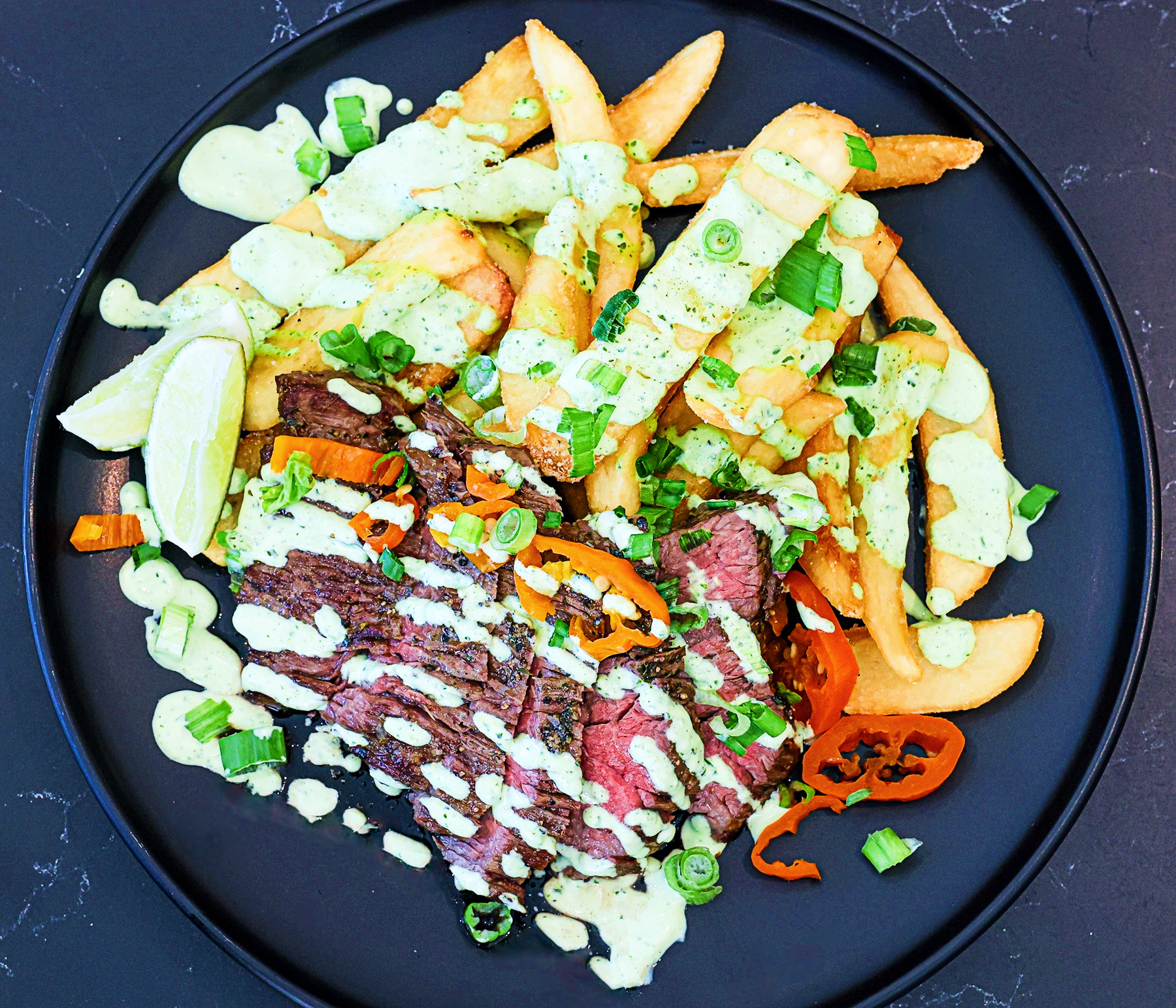 A plate of steak and fries topped with creamy green sauce, chopped herbs, peppers, and cucumber slices, finished with lime wedges.