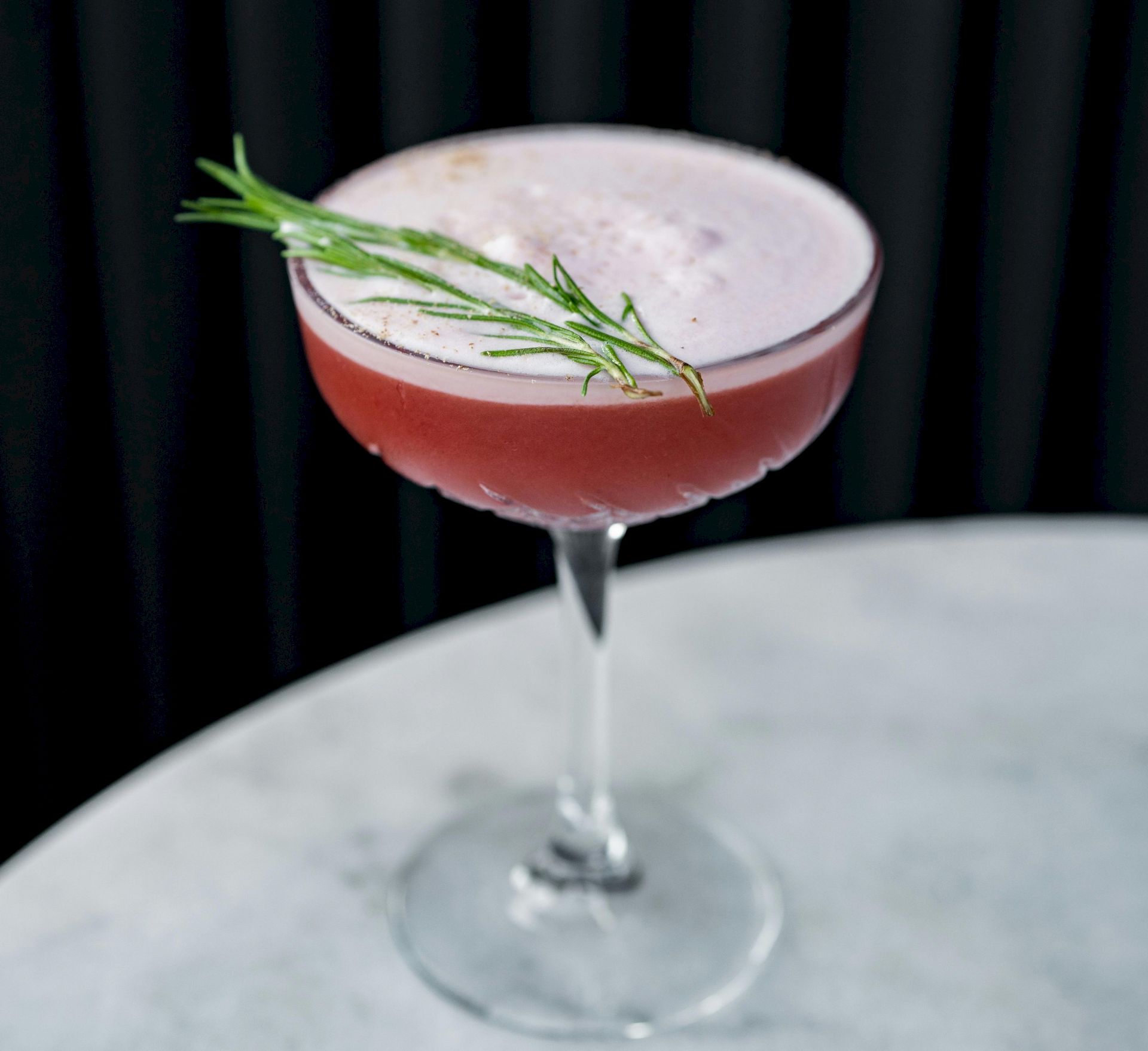 A pink cocktail in a coupe glass, garnished with fresh rosemary sprigs, sitting against a dark background.