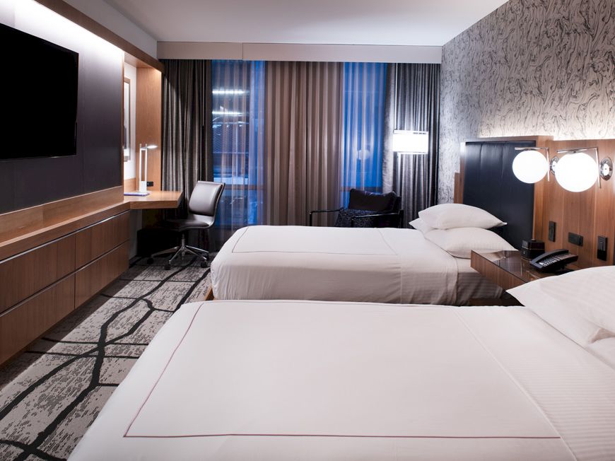 A modern hotel room with two neatly made beds, bedside lamps, a TV, and large curtains by a window.