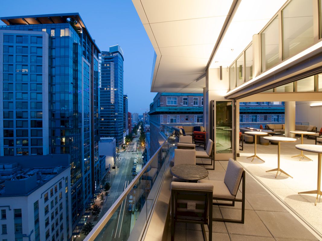 A modern high-rise balcony overlooks a downtown street with outdoor lounge seating and skyscrapers at blue hour.